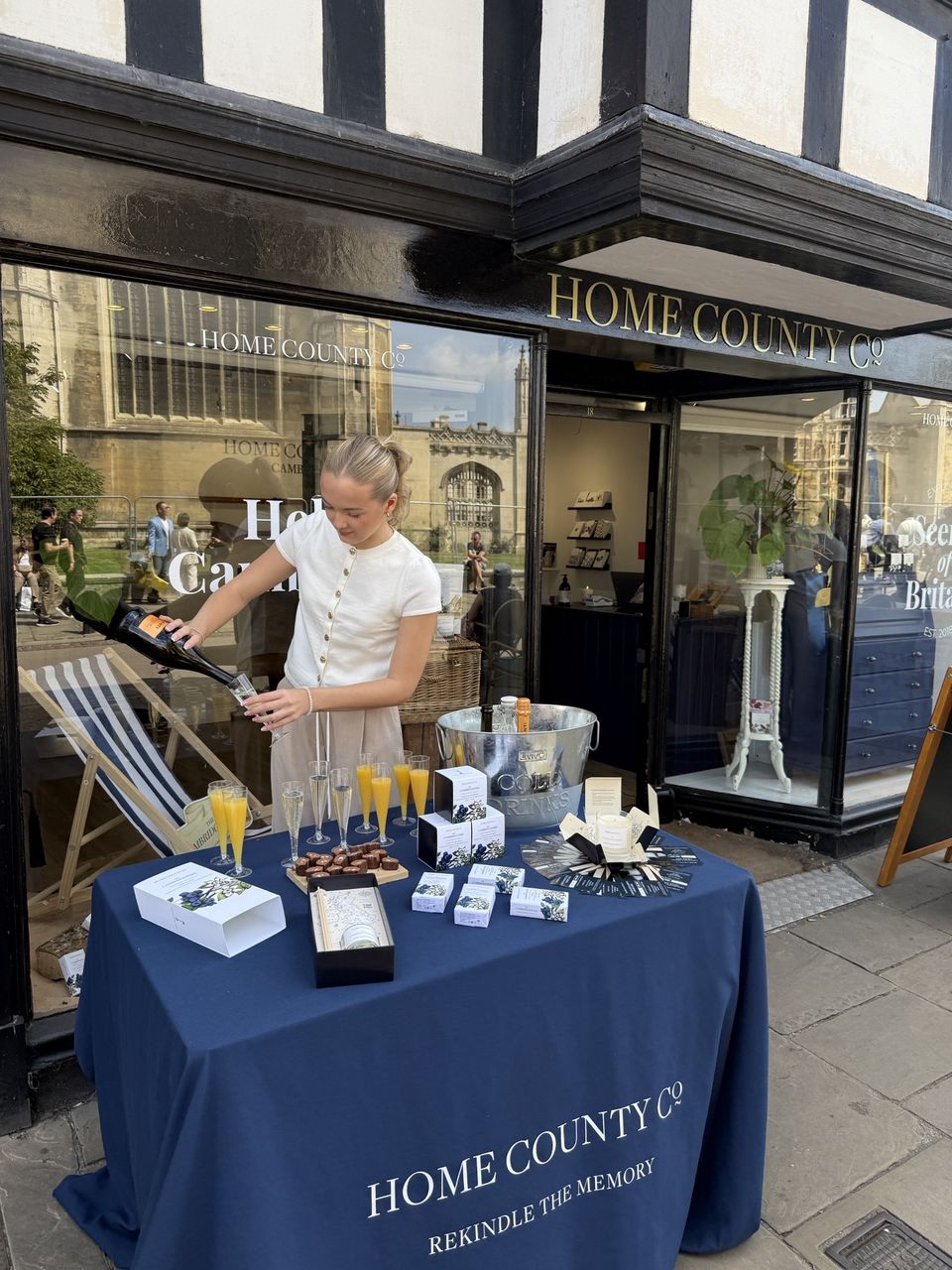 Me pouring a glass of bubbly at the graduates' table outside the Cambridge shop, with the Cambridgeshire candles and chocolates laid out