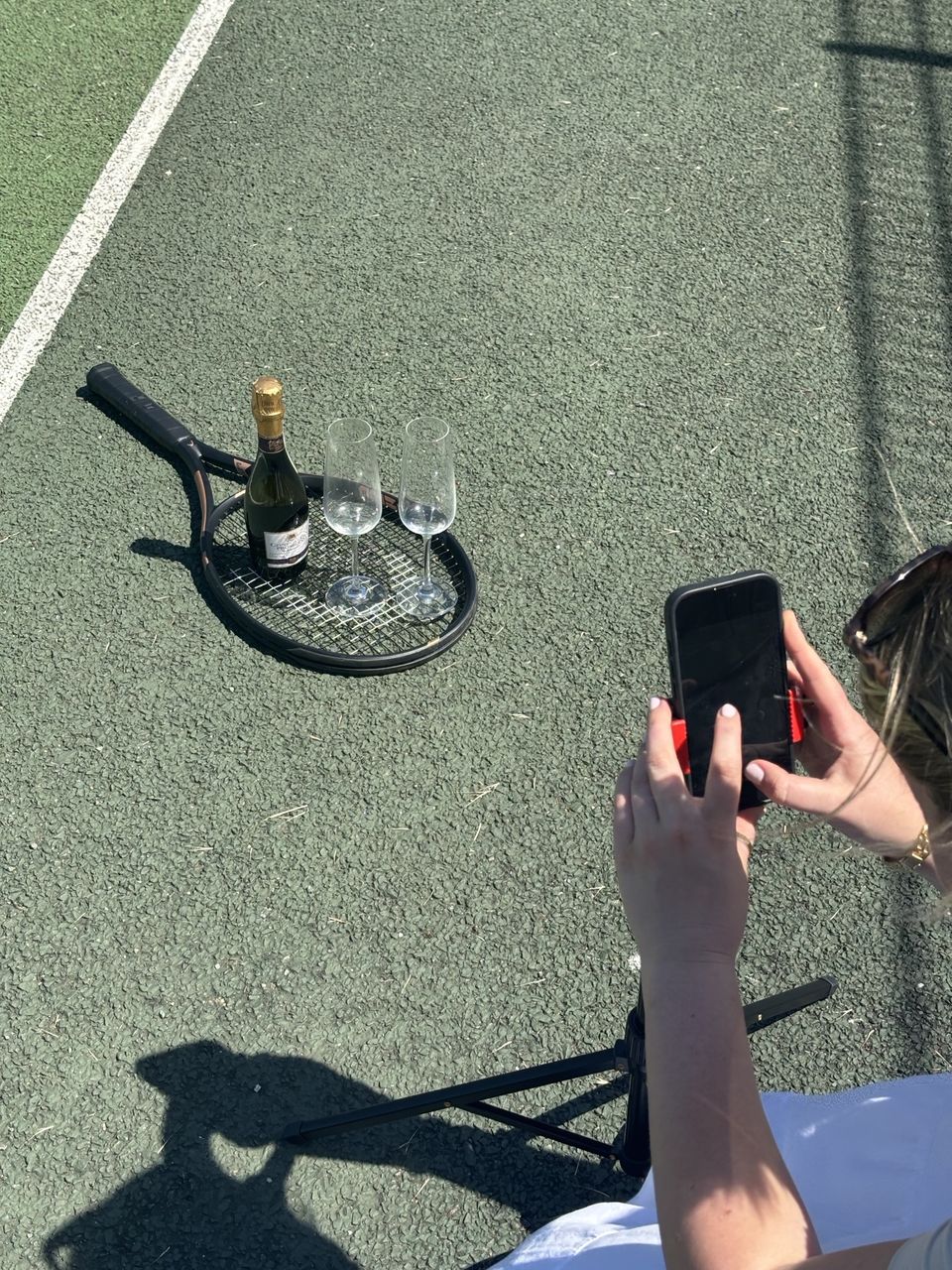 Behind the scenes on the tennis court: a bottle of fizz and two glasses on a racket, with me framing the shot on my phone