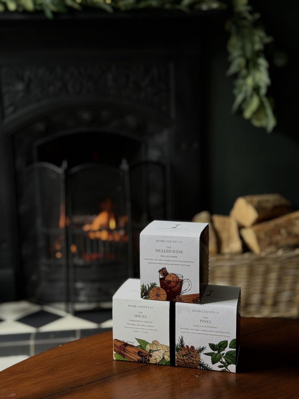 The three Winter Collection boxes, The Mulled Wine, The Spices and The Pines, stacked on a table in front of a lit fire