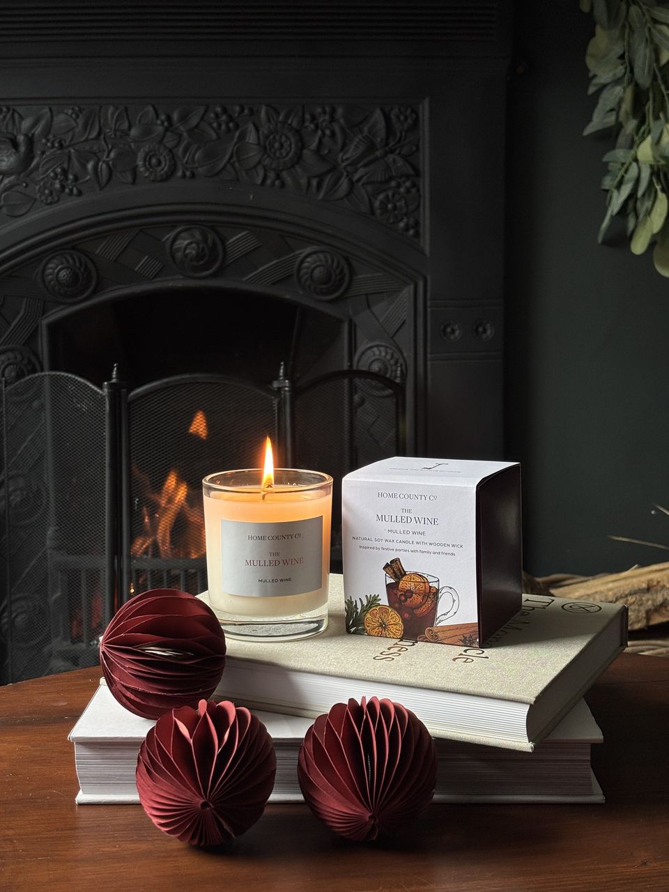 The Mulled Wine candle lit beside its box on a stack of books, with deep red paper baubles and a fire behind